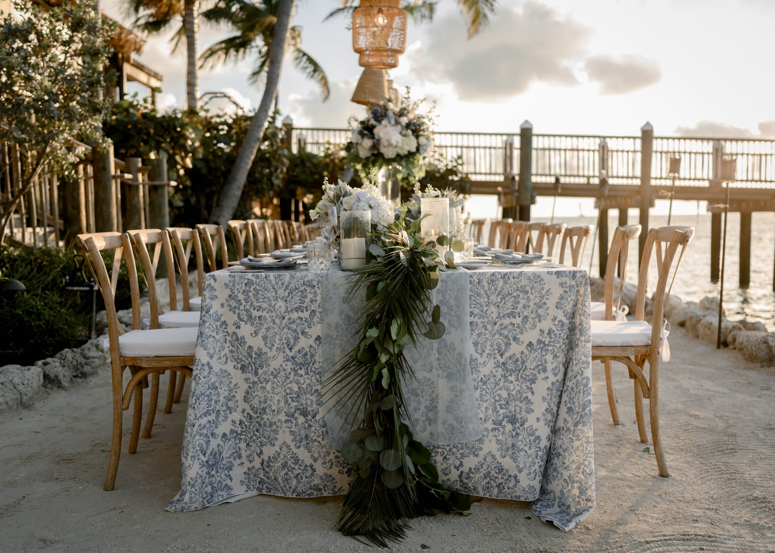 A dinner table set up on the beach with a blue and white table cloth.