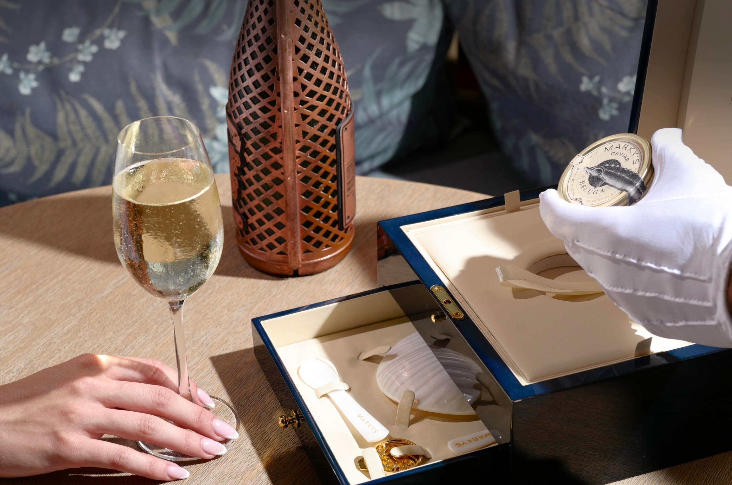 A woman holding a glass of wine, while a white gloved hand holds a jar of caviar.
