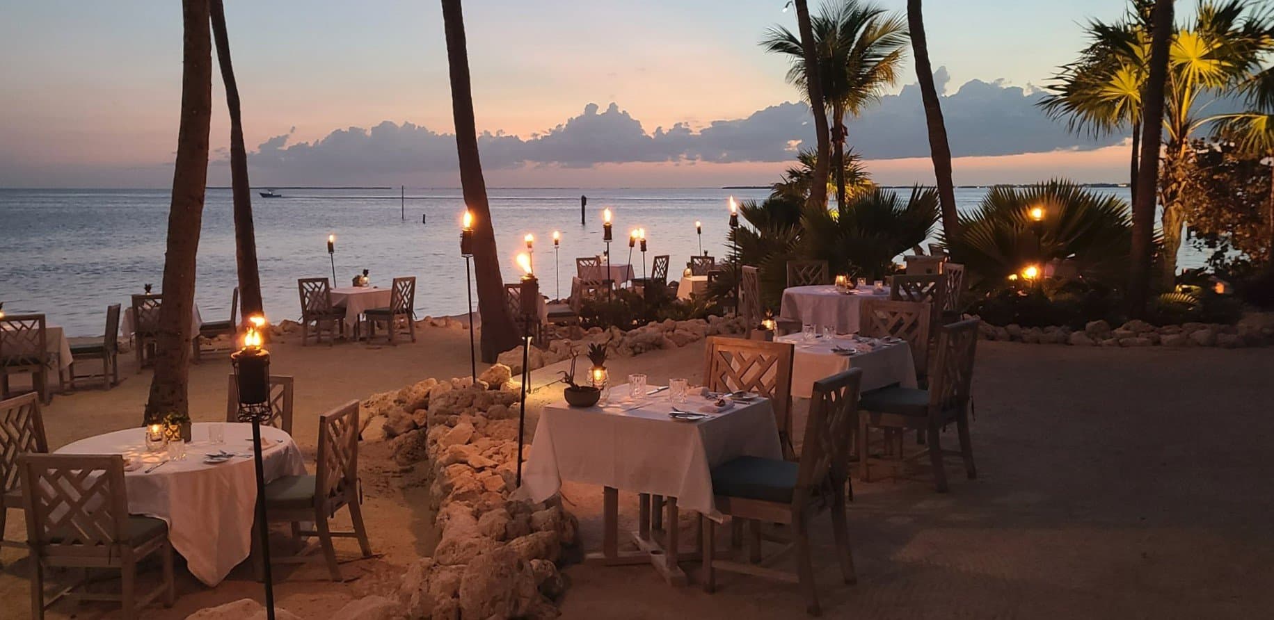 A row of dinner tables set with white table cloths on the beach with tiki torches lit around them.