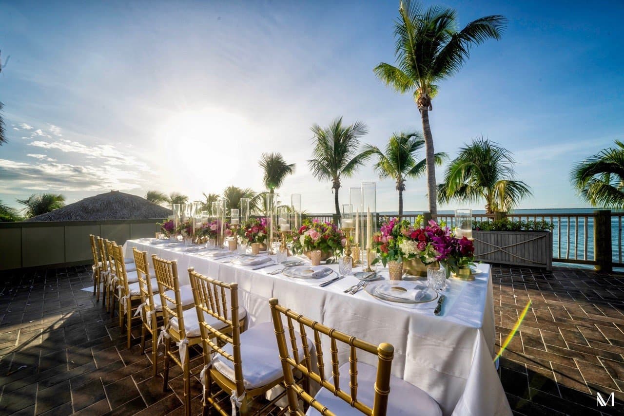 Image a table set with flowers and a white table cloth outside with palm trees in the distance.