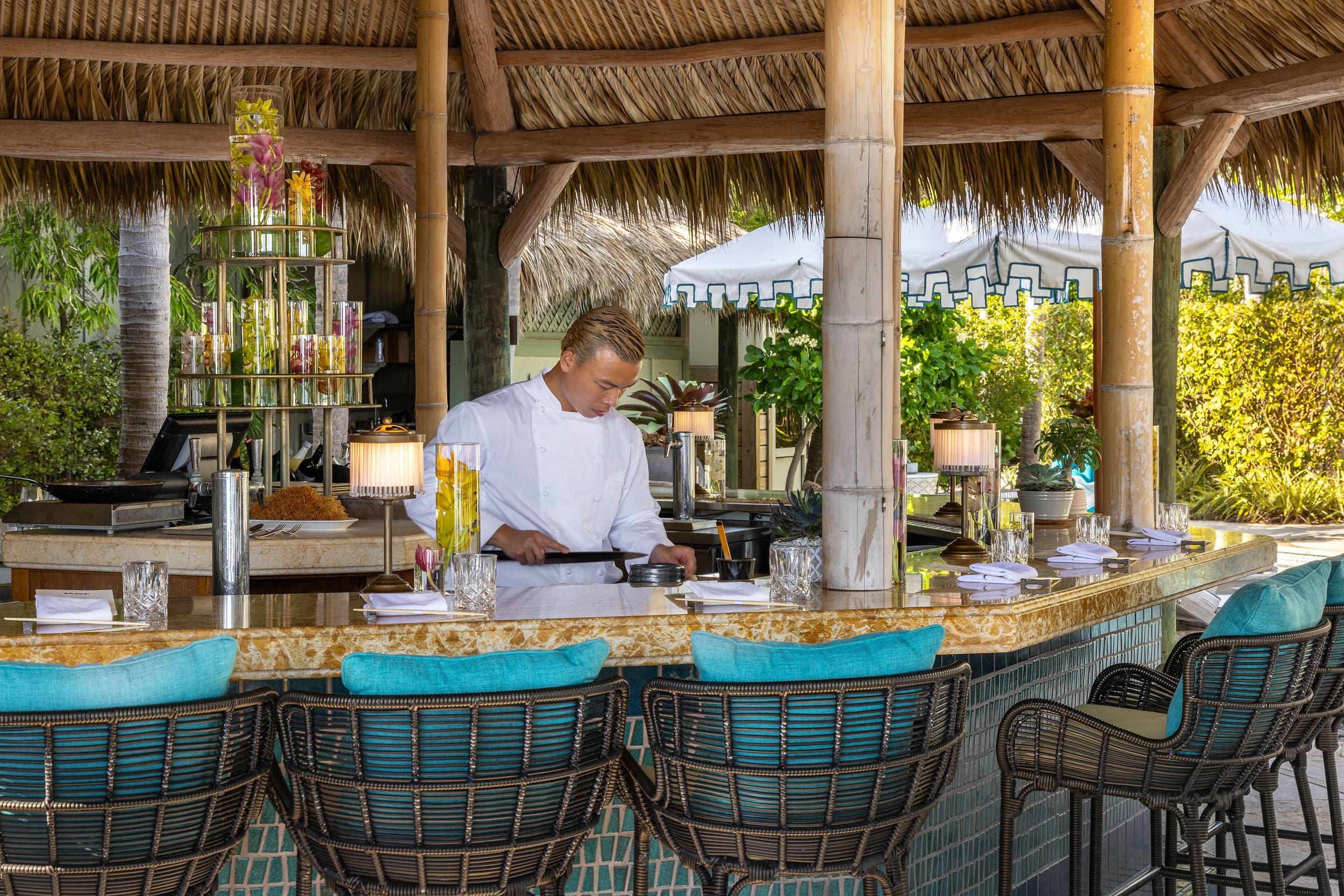 Chef preparing a dish at the thatched-roof chef’s counter at Little Palm Island Resort & Spa in the Florida Keys, with turquoise-cushioned wicker bar stools and tropical gardens behind.