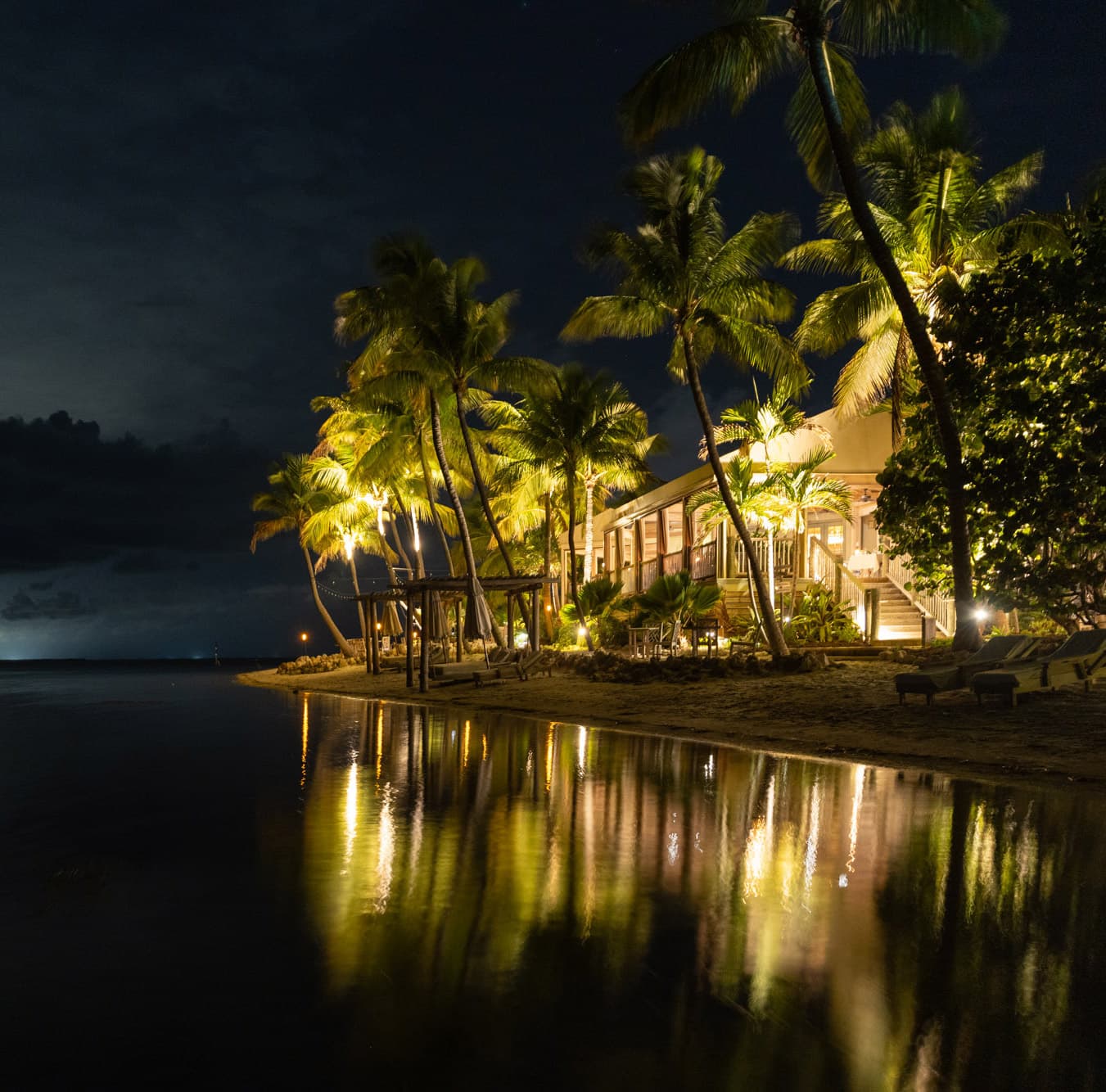 Uplit palm trees and a beachfront guest building reflected in still water at night at Little Palm Island Resort in the Florida Keys