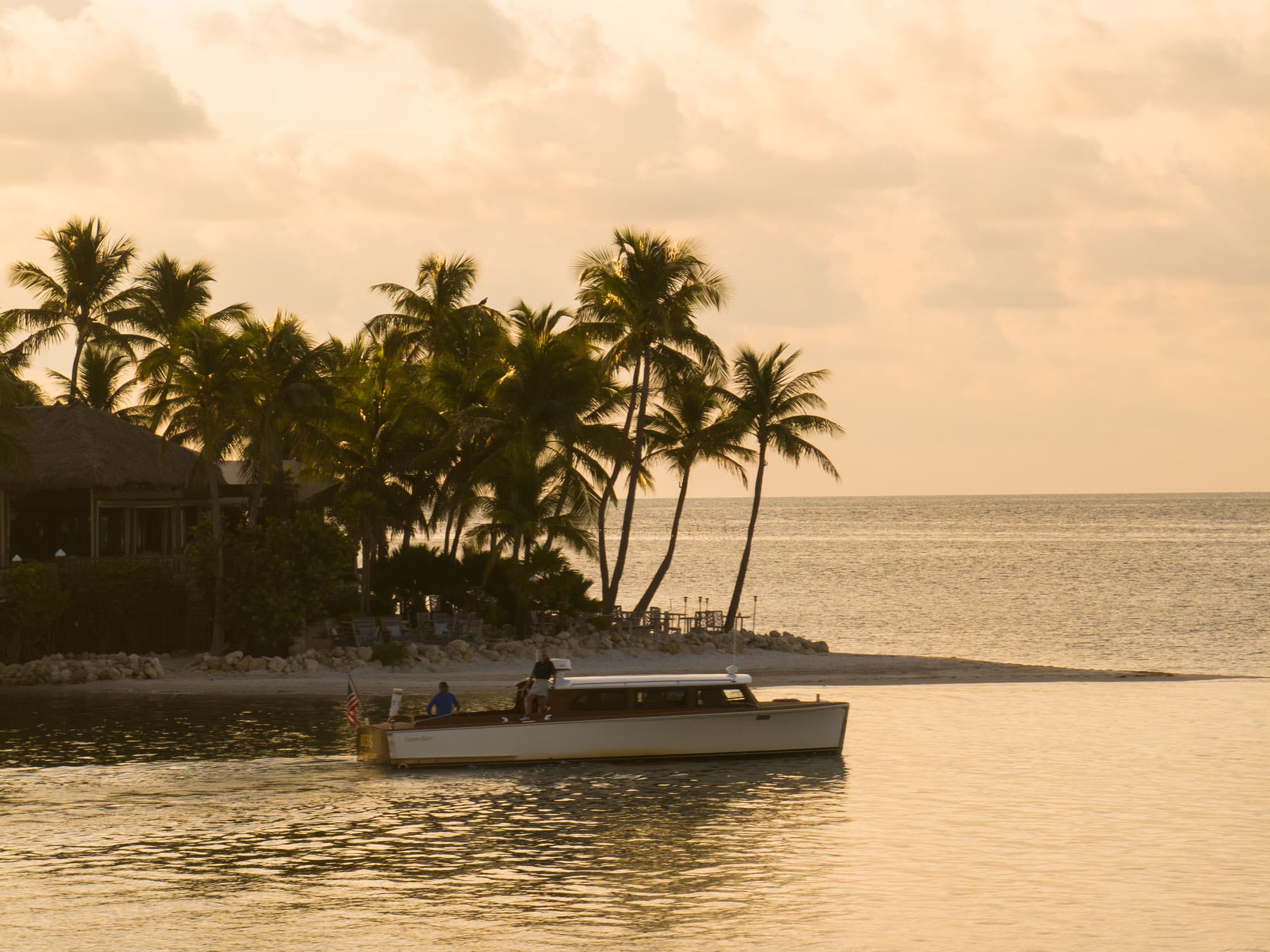 Vintage motor launch departing Little Palm Island Resort & Spa at golden hour, framed by palm trees and thatched-roof bungalows in the Florida Keys.