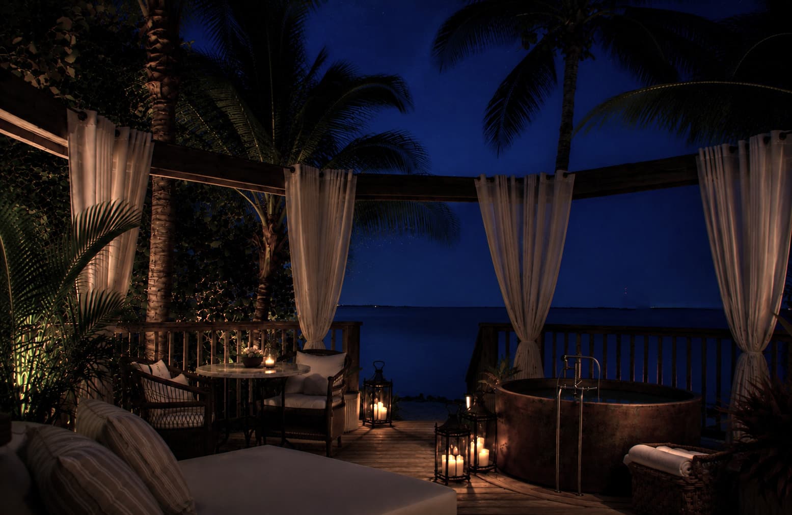 Private candlelit soaking tub on a tropical oceanfront deck at night, framed by palms and white linen curtains