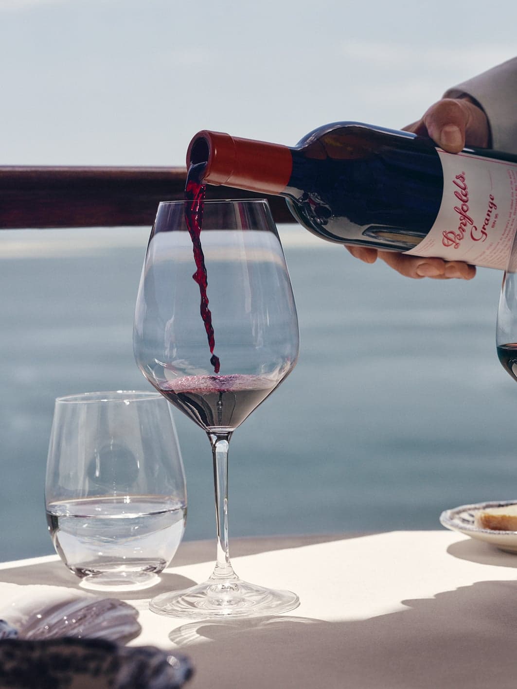 Server pouring Penfolds Grange red wine into a glass at an oceanfront dining table overlooking calm blue water.